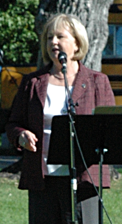 A photograph of a blonde woman wearing a white shirt, a purple jacket, and black dress pants standing in front of a black microphone